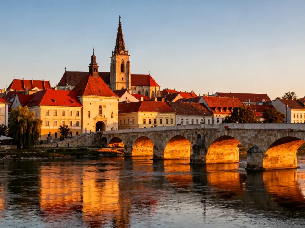 Panoramiczny widok na centrum Mariboru o zachodzie słońca z historycznym mostem nad rzeką Drawą i zabudową starego miasta.