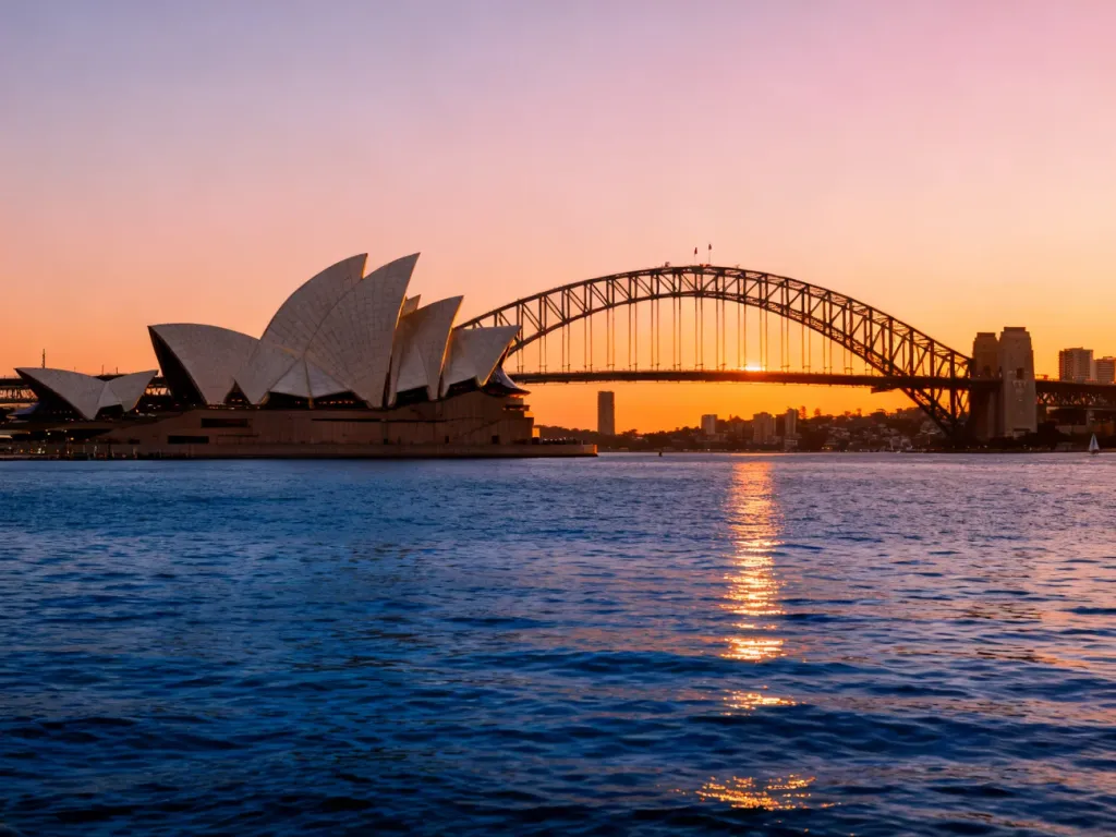 Widok na Operę w Sydney i Harbour Bridge o zachodzie słońca, odbijające się w spokojnych wodach zatoki.